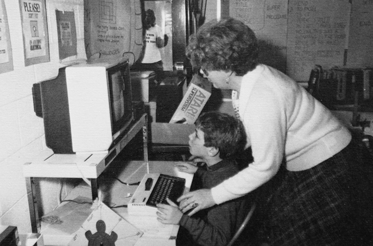 Black and white photograph of a teacher and a student at a computer workstation.